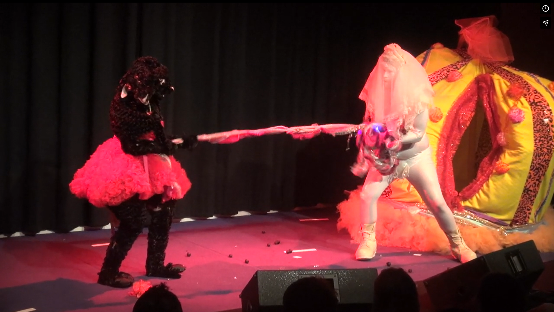 Two costumed performers on stage, one in a black fluffy suit and red tutu, the other in a white bodysuit and veil, tugging a 